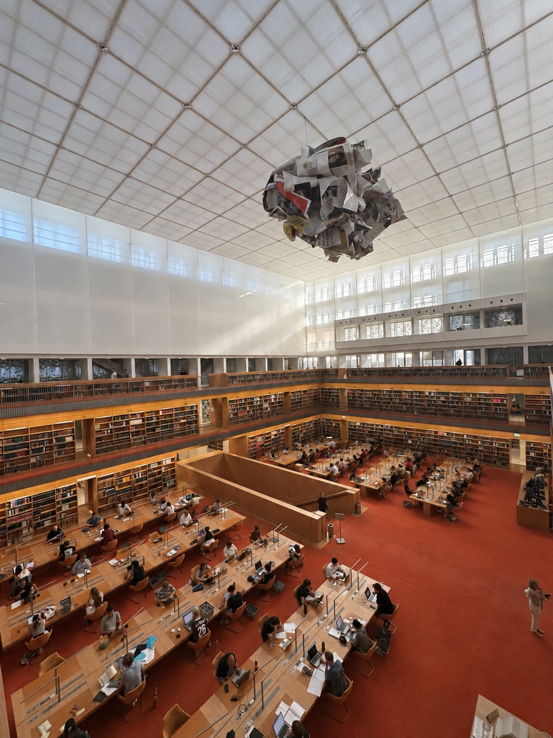 A spacious library interior in Berlin with rows of bookshelves, reading tables, a geometric ceiling and a suspended newspaper sculpture.