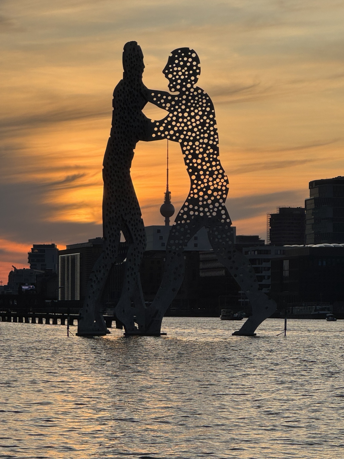 Molecule Man sculptures silhouetted against a vivid sunset over the Spree in Berlin, Germany, with the Fernsehturm visible between the figures.
