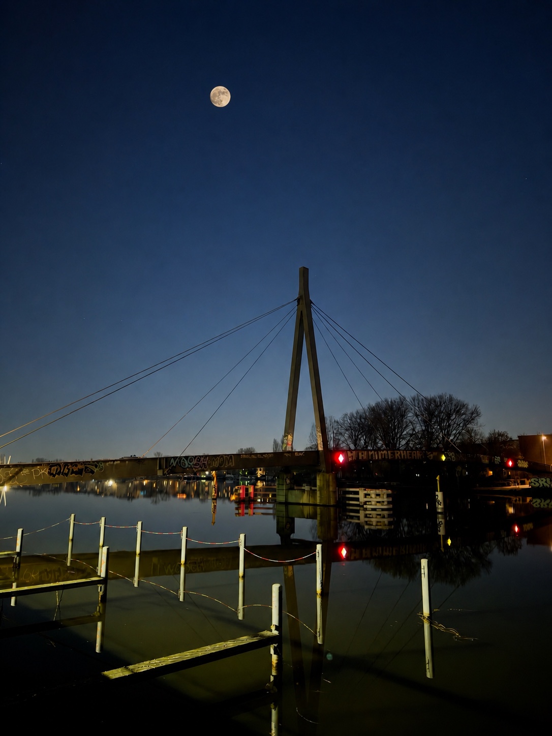 A full moon above a cable-stayed bridge over calm water in Berlin, Germany, with graffiti-covered railings, reflections and moored structures in the foreground.