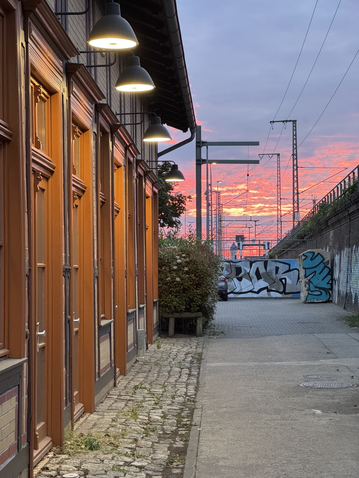 A railway-side street in Berlin at dusk, with warm exterior lamps, historic building facades, railway wires and a vivid pink sunset beyond graffiti-covered walls.