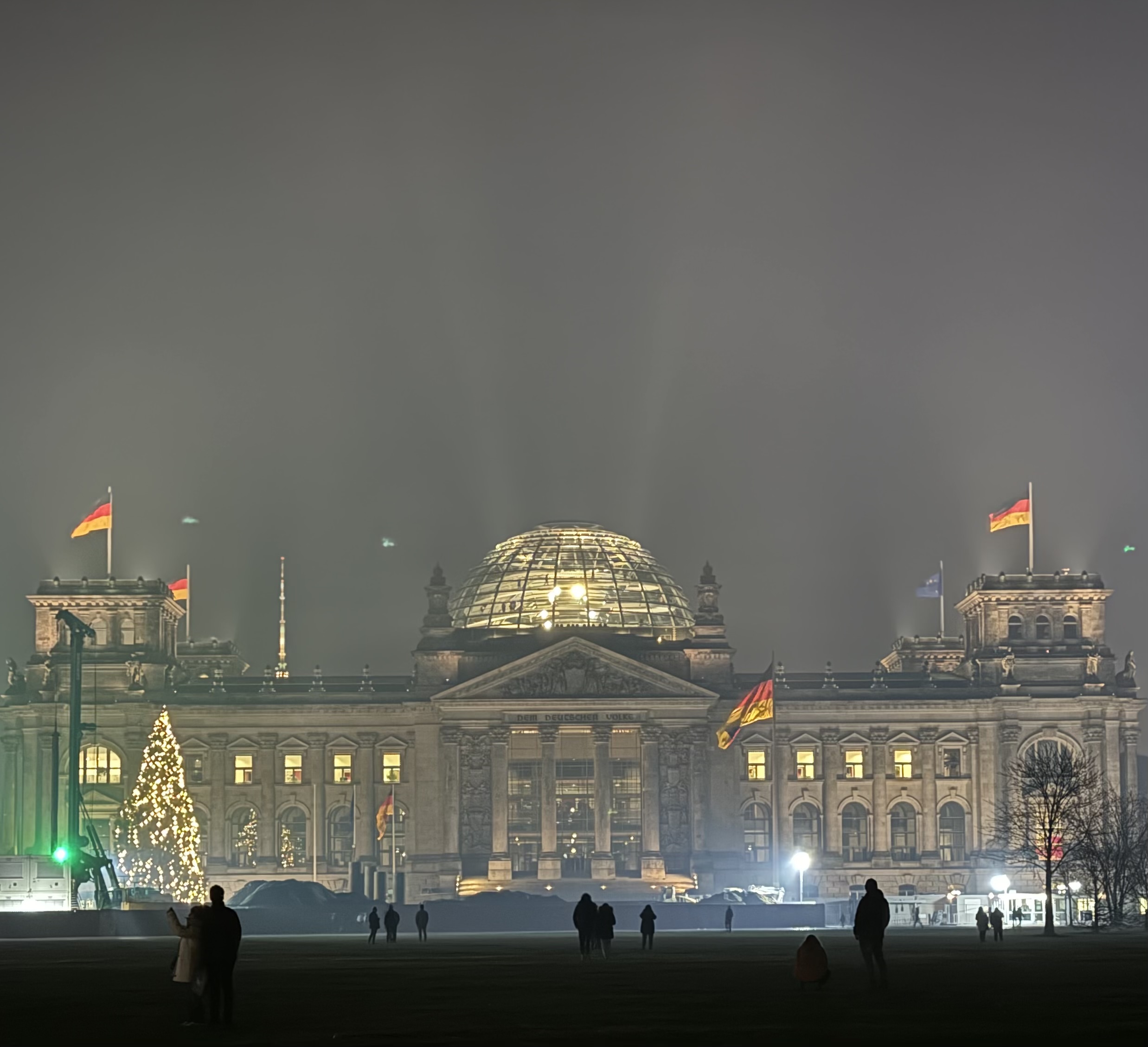 The Reichstag building in Berlin at night, illuminated through winter fog with German flags, a Christmas tree and people in the foreground.
