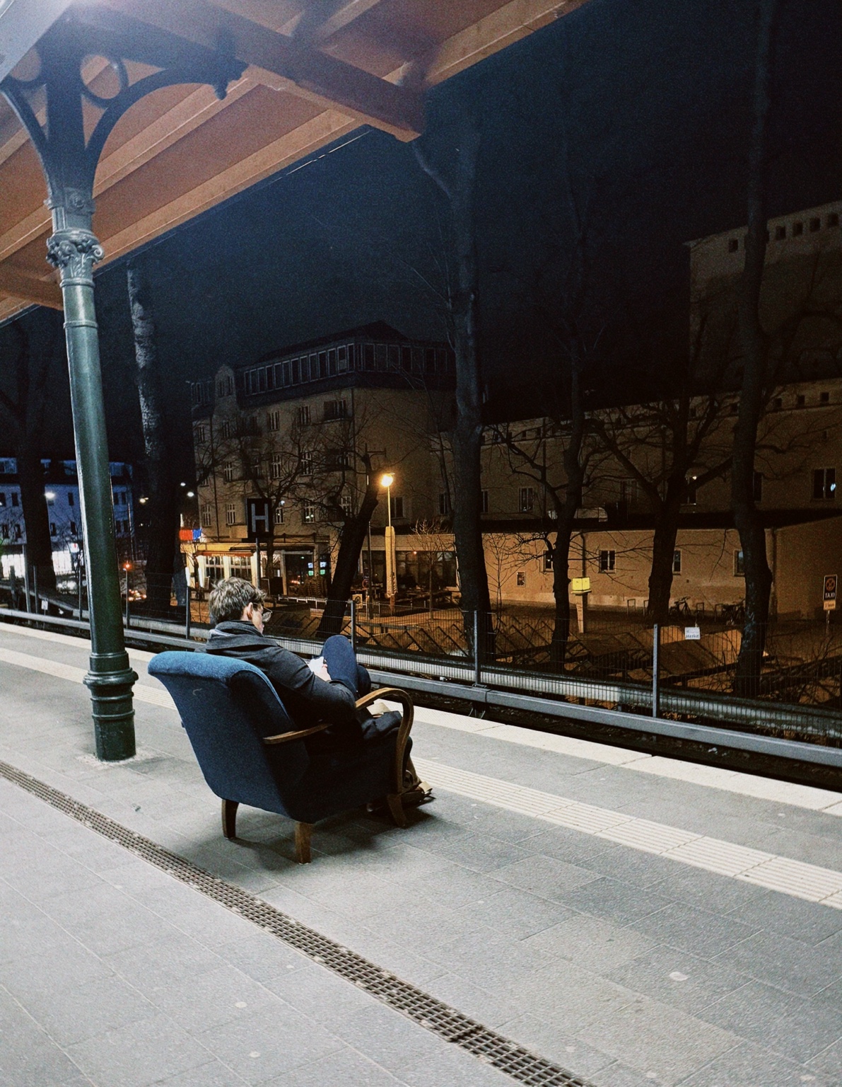 A lone person sitting in a blue armchair on a Berlin railway station platform at night, with historic buildings and warm street lights beyond the tracks.