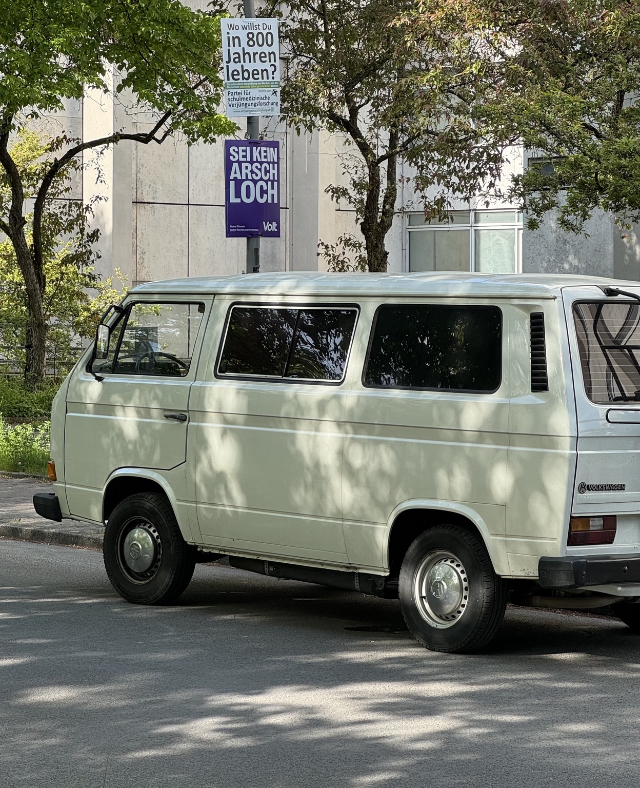 A vintage Volkswagen van parked on a Berlin street beneath trees, with political campaign posters visible behind it.