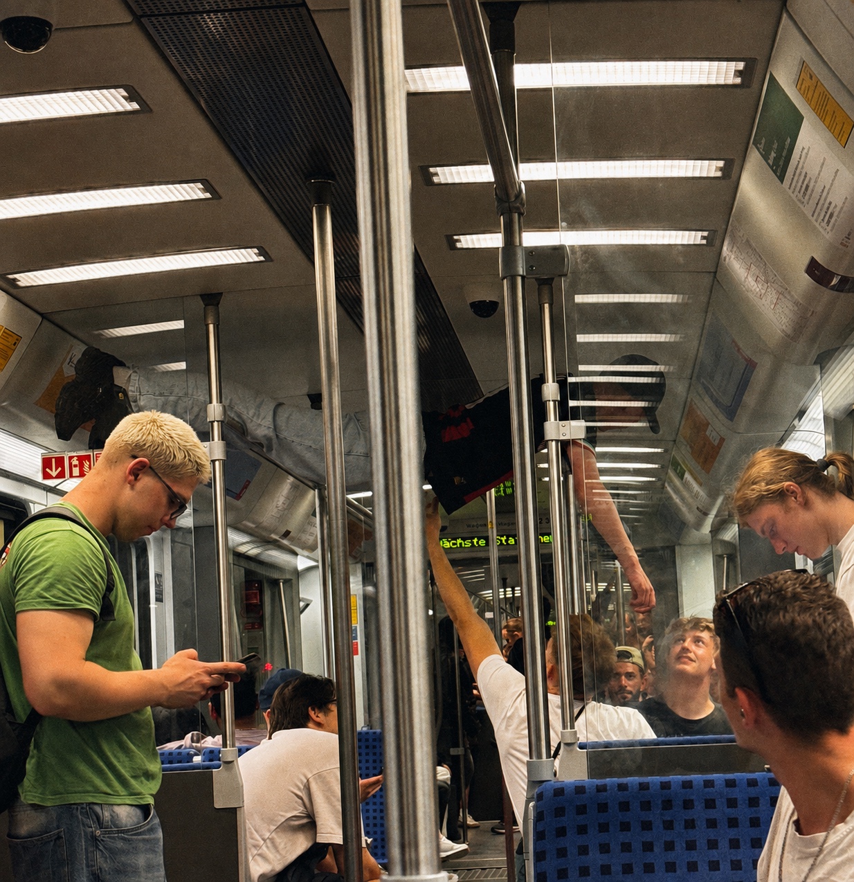 Passengers inside a Berlin train near Warschauer Straße station, with reflections, handrails and a digital station display visible through the carriage.