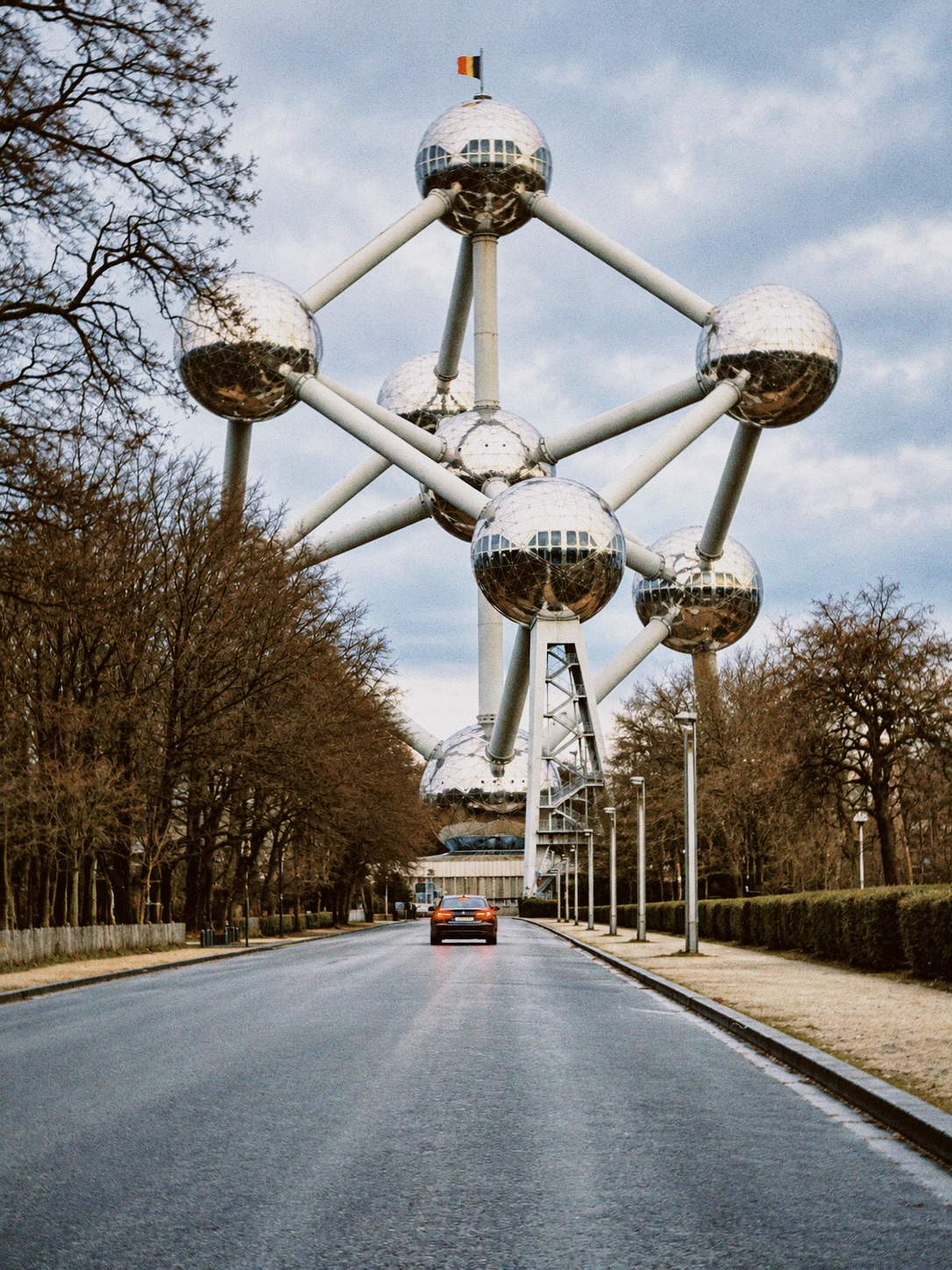 The Atomium in Brussels rising above a tree-lined avenue, with its steel spheres reflecting the winter sky and a car on the road below.