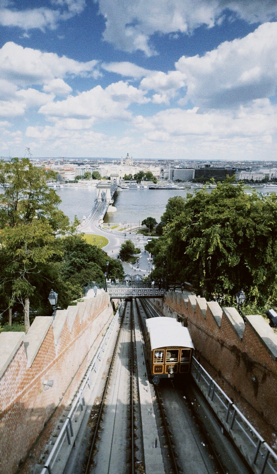 The Buda Castle Funicular descending toward the Danube in Budapest, Hungary, with the Chain Bridge, river and city skyline beyond under a dramatic cloud-filled sky.