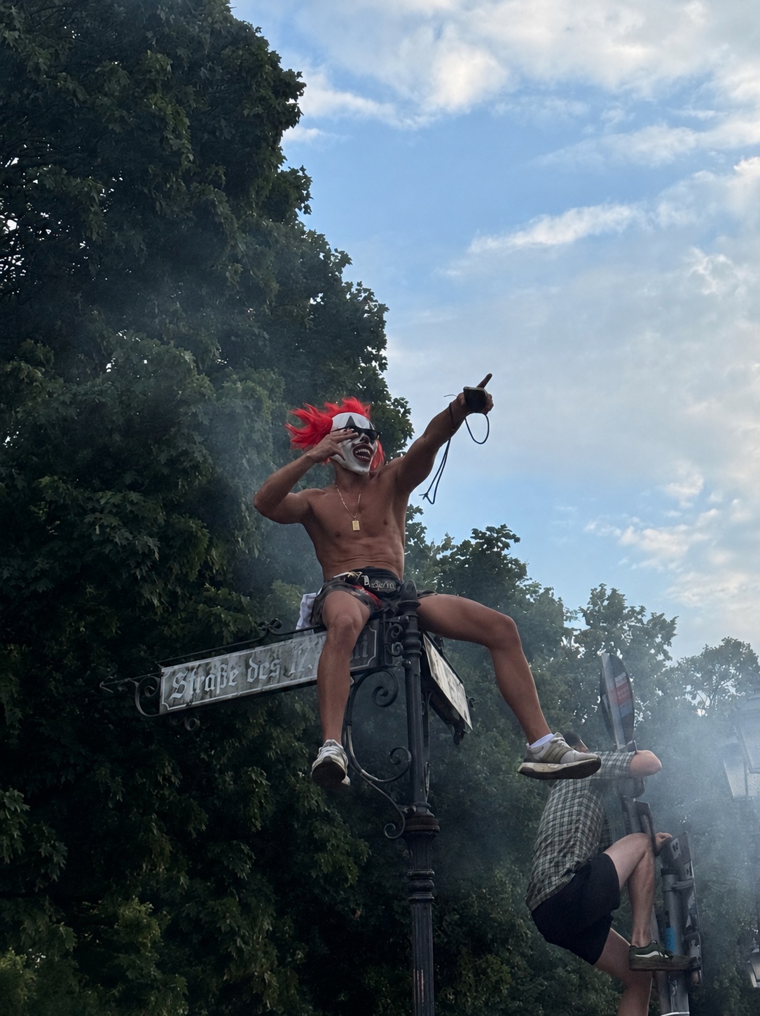 A masked performer with bright red hair climbing above a Berlin street during a public event, photographed against trees and a smoky summer sky.