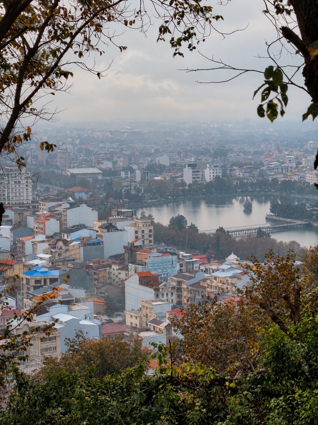 A misty panoramic view over a densely built city in Gilan, Iran, with a calm lake, bridge and autumn trees in the foreground.