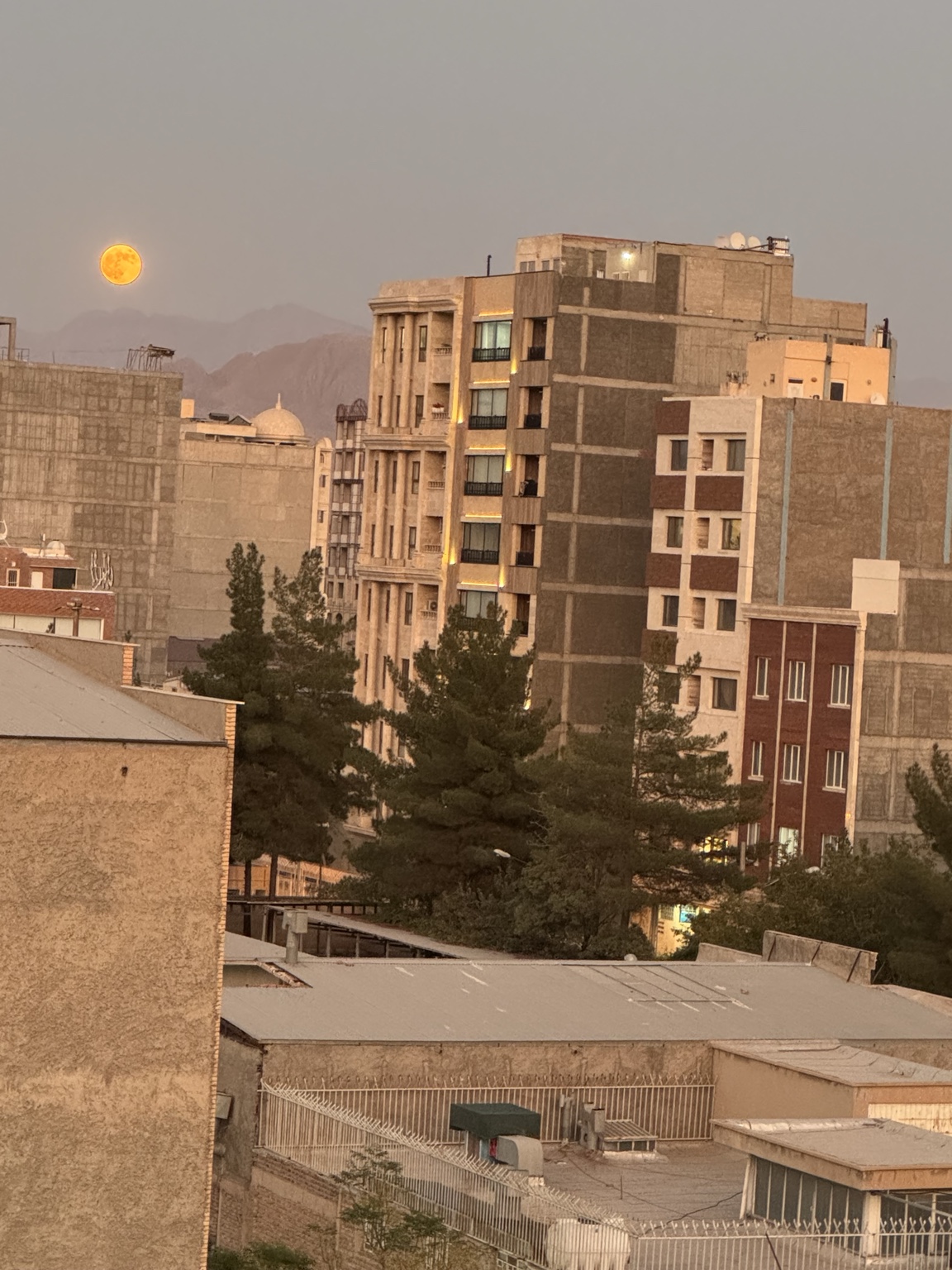 A full moon rising over the rooftops of Kerman, Iran, with mountains and residential buildings in the evening light.