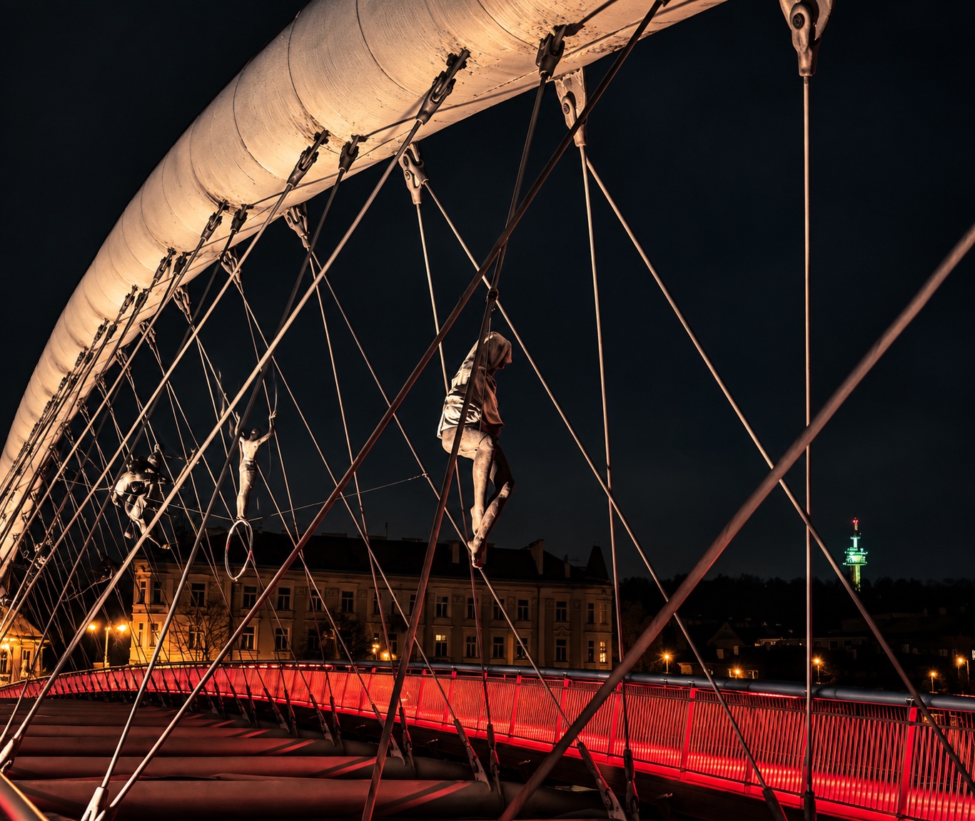 A suspension footbridge illuminated at night in Kraków, Poland, with suspended human figures, a red-lit railing and city buildings beyond.