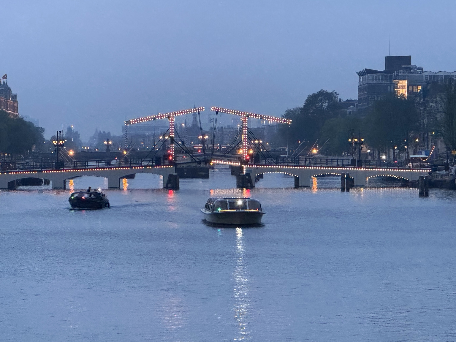 The illuminated Magere Brug over the Amstel in Amsterdam, Netherlands, at blue hour, with boats on the river and city buildings beyond.