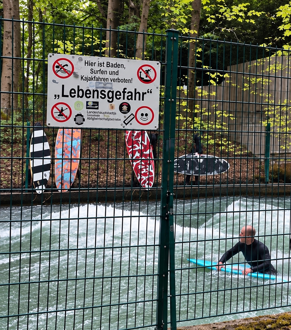 A surfer in a wetsuit riding a board in the fast-flowing Eisbach in Munich, with surfboards behind a fence and a German warning sign reading ‘Lebensgefahr’.