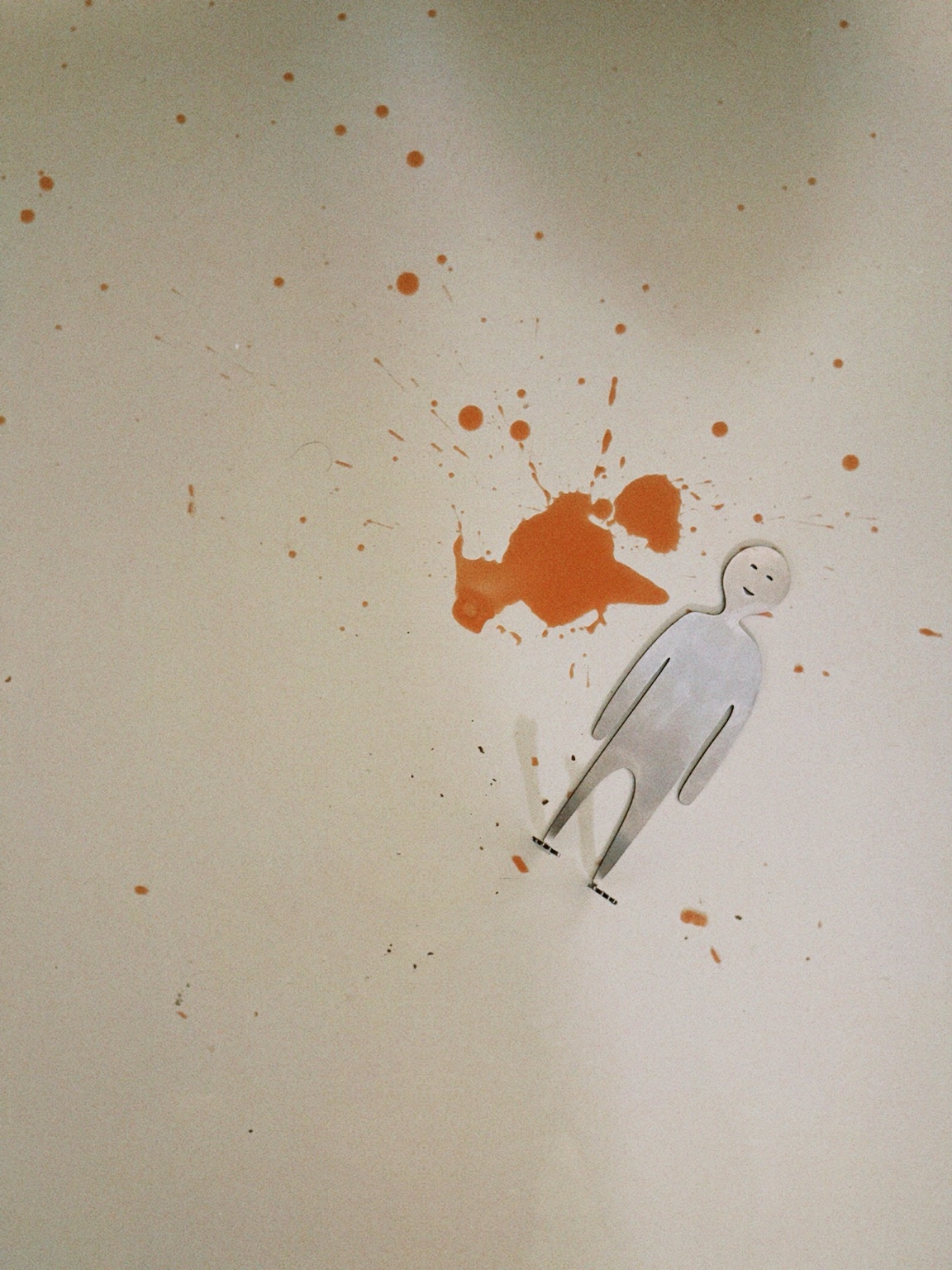 A minimalist white human figure beside an orange paint-like splash on a pale wall, photographed in Berlin, Germany.